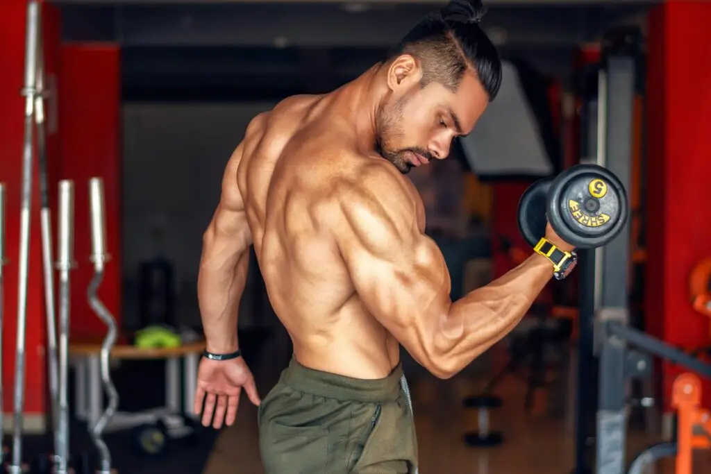A fit, shirtless man showcasing biceps while lifting a dumbbell in a gym setting.